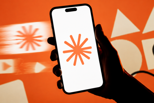 Mão segura um smartphone com o símbolo do Claude sobre fundo laranja, enquanto formas de conteúdo passam em movimento, sugerindo criação rápida para redes sociais.